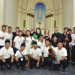 Group of young musicians and adults posing in a church, holding violins and a cello, with a statue in the background.