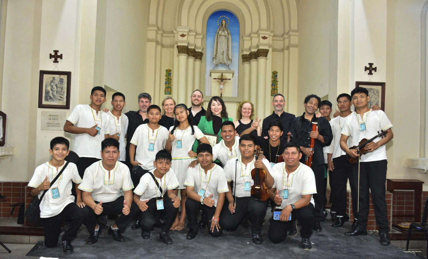 Group of young musicians and adults posing in a church, holding violins and a cello, with a statue in the background.