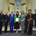 Group of nine people in formal attire posing with a framed certificate inside a church sanctuary with an altar in the background.