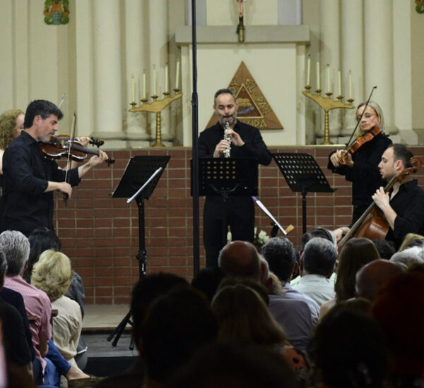 Musicians performing a small classical ensemble (violin, clarinet, cello) on a church stage with audience.