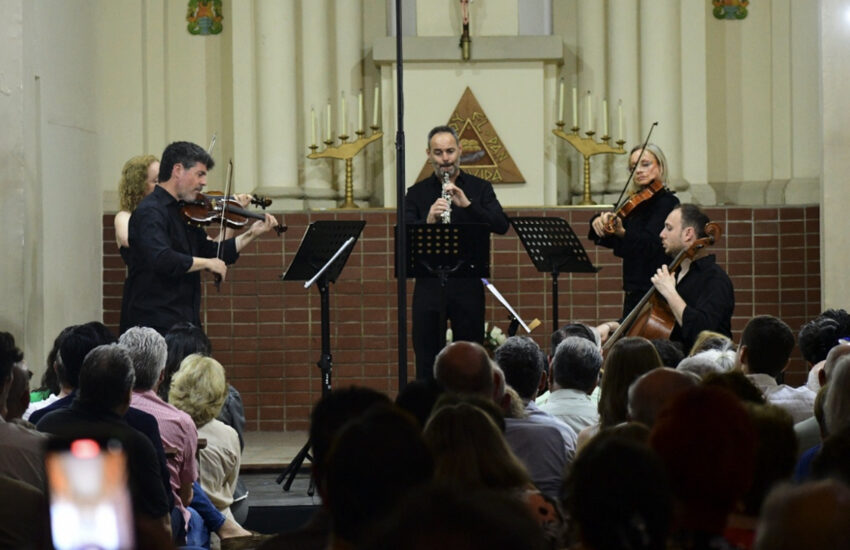 Musicians performing a small classical ensemble (violin, clarinet, cello) on a church stage with audience.