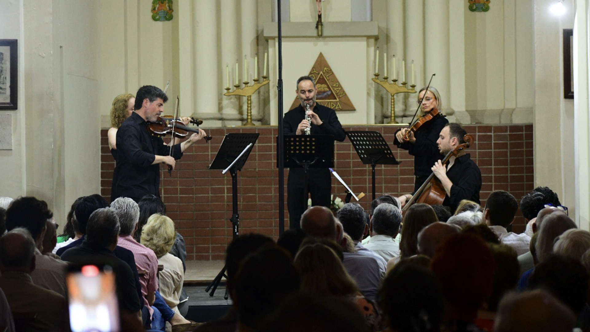 Musicians performing a small classical ensemble (violin, clarinet, cello) on a church stage with audience.