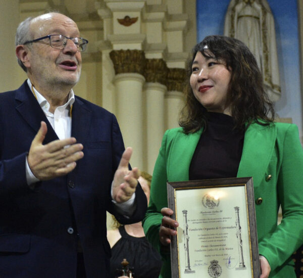 Woman in a bright green blazer holds a framed certificate as a man in a dark suit gestures and speaks beside her at an award ceremony.