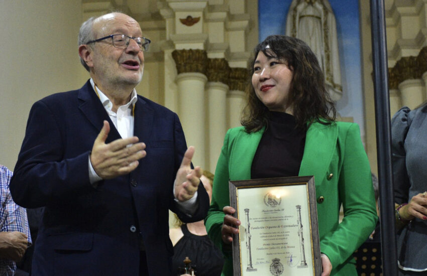 Woman in a bright green blazer holds a framed certificate as a man in a dark suit gestures and speaks beside her at an award ceremony.