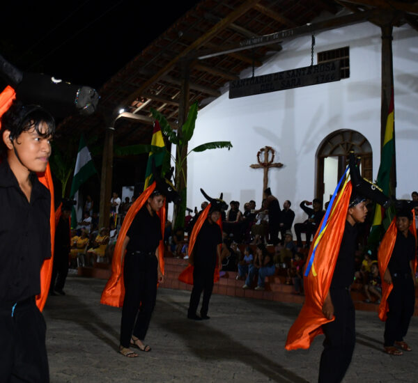 Nighttime outdoor dance performance with performers in black and bright orange capes, audience seated on steps nearby.