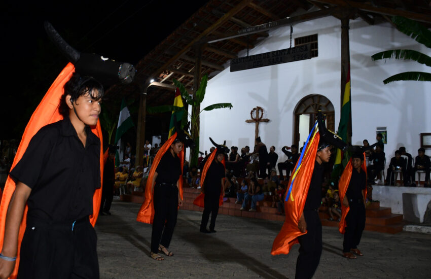 Nighttime outdoor dance performance with performers in black and bright orange capes, audience seated on steps nearby.