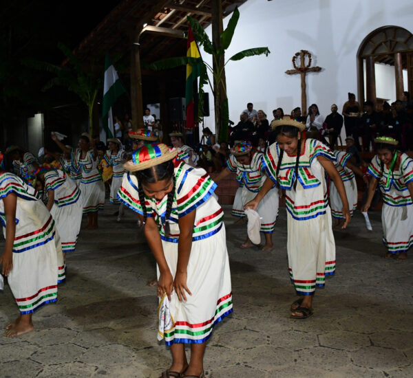 Group of children in white striped dresses dance at night, with spectators watching from the steps behind them.