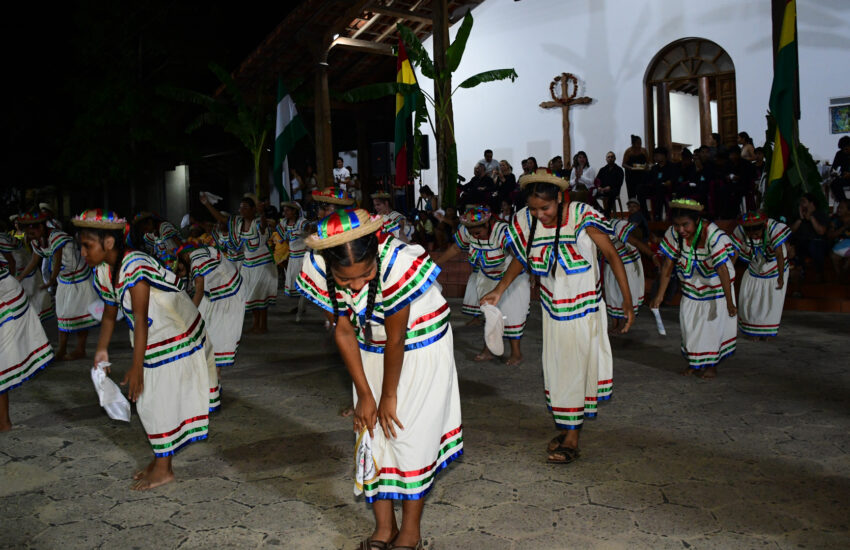 Group of children in white striped dresses dance at night, with spectators watching from the steps behind them.