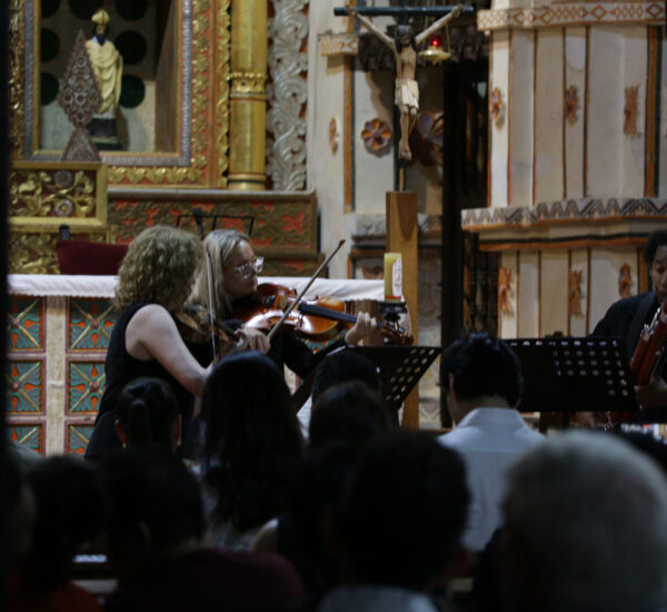 Musicians perform in a richly decorated church: two violinists and a bassoonist with audience watching.