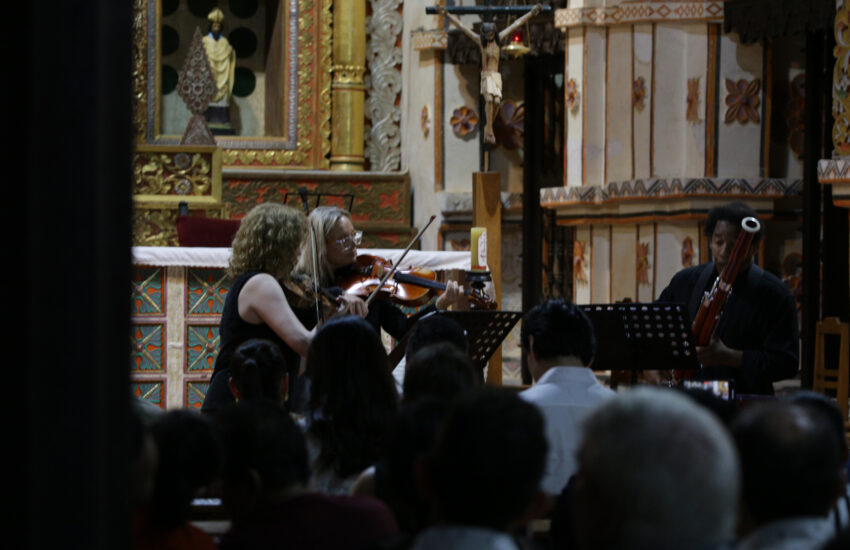 Musicians perform in a richly decorated church: two violinists and a bassoonist with audience watching.