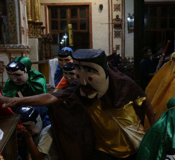 Masked performers in colorful robes and large carved masks perform indoors while a small audience watches.
