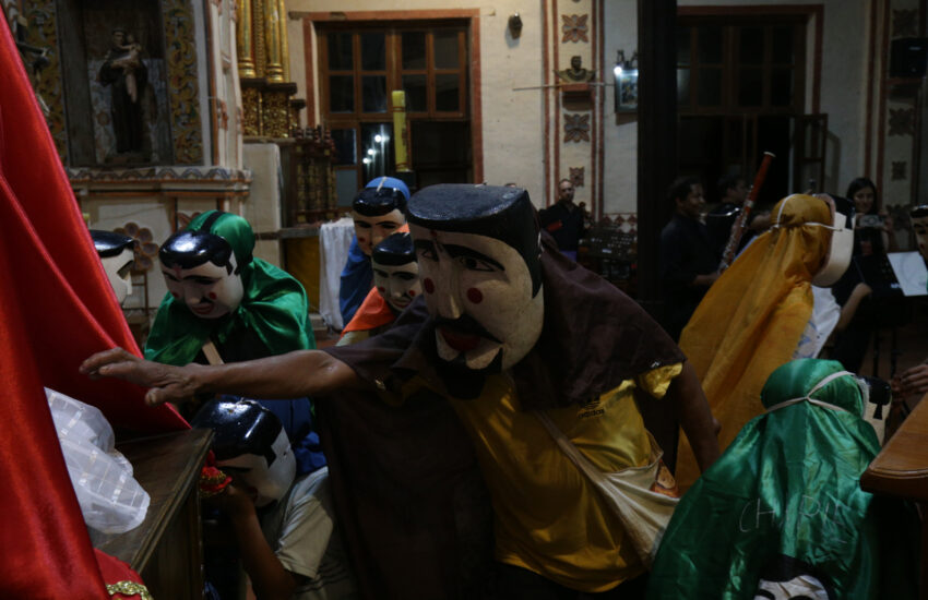 Masked performers in colorful robes and large carved masks perform indoors while a small audience watches.