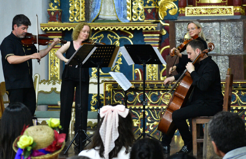 String quartet performing on a stage in a richly gilded, ornate hall with audience watching in the foreground.