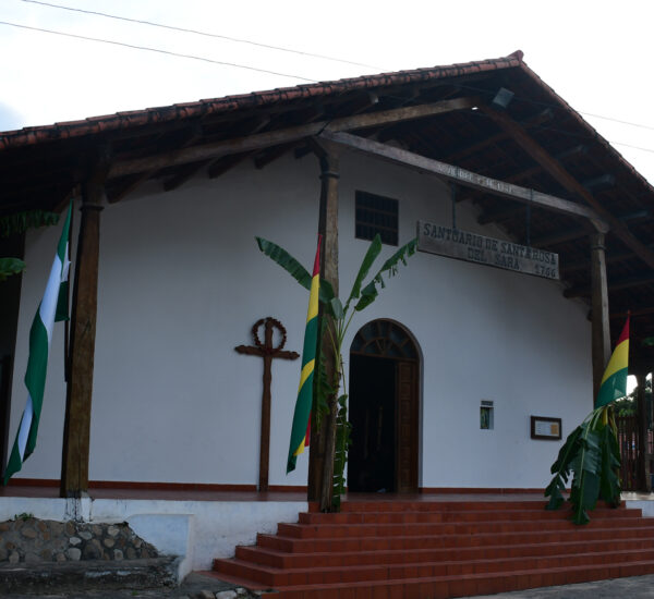 White church with a red-tiled roof and wooden porch, flanked by banana plants and flag banners outside the entrance. Cross mounted on wall beside door.