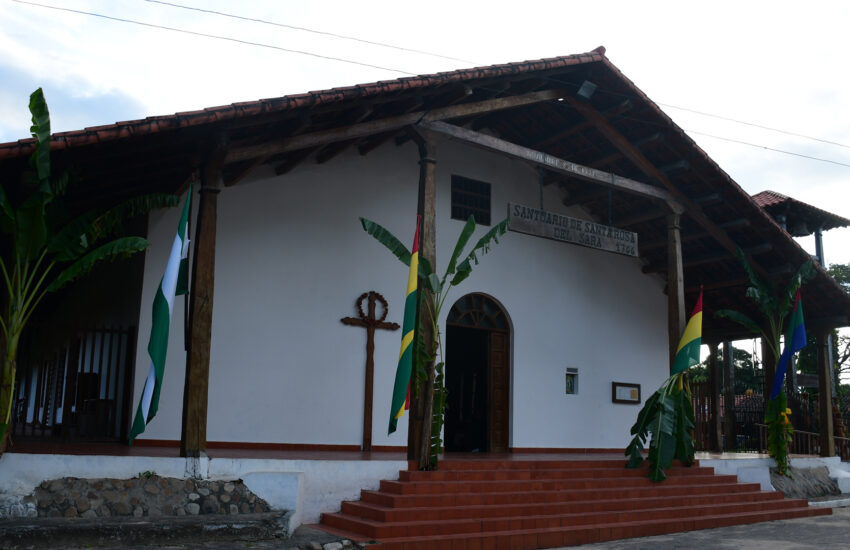 White church with a red-tiled roof and wooden porch, flanked by banana plants and flag banners outside the entrance. Cross mounted on wall beside door.