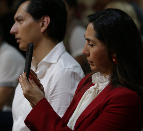 Woman in red blazer and white blouse looking down at a black smartphone held vertically in her hands in a dim room.