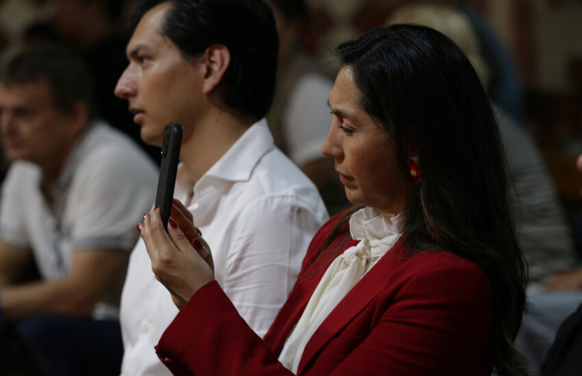 Woman in red blazer and white blouse looking down at a black smartphone held vertically in her hands in a dim room.