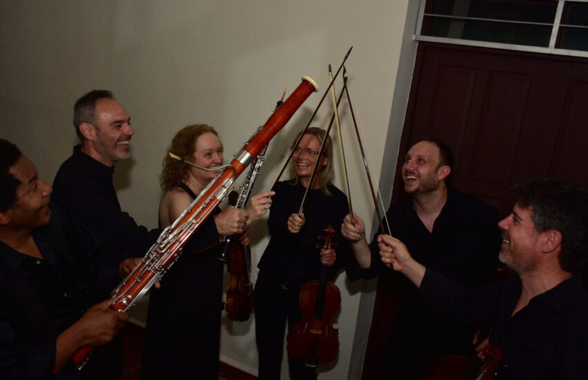 Six musicians in black stand in a circle, smiling, each holding bows or string instruments (violin/viola) and a bassoon at a casual group moment.