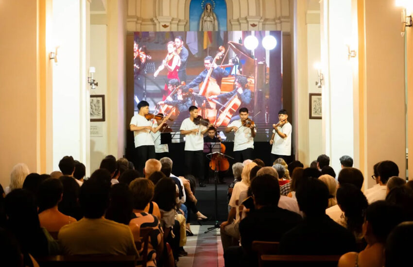A string quartet performing on a stage inside a church, with an audience seated in front and a large screen behind them showing the performance.
