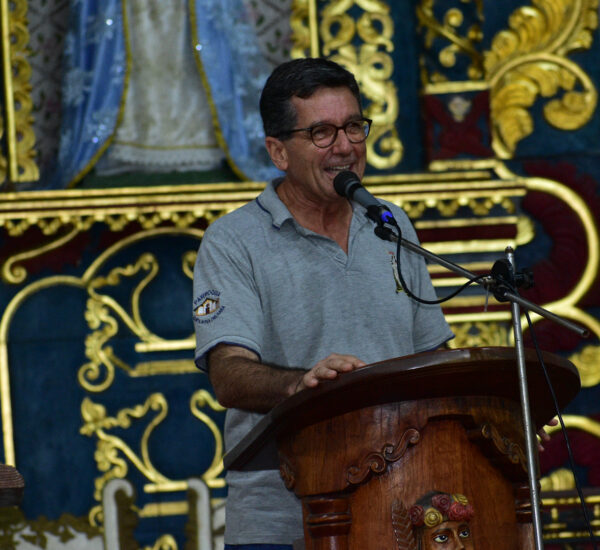 Man speaking into a microphone at a carved wooden podium, with an ornate gold-and-blue backdrop behind him.