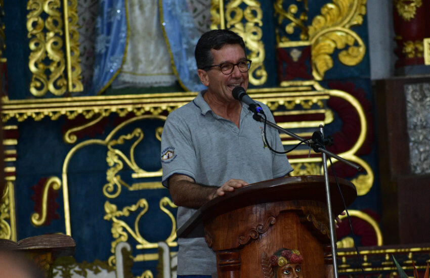 Man speaking into a microphone at a carved wooden podium, with an ornate gold-and-blue backdrop behind him.