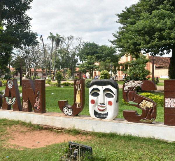 Decorative outdoor sculpture spelling a word with large art-painted letters in a park; each letter features masks and figures, with trees and brick buildings in the background.