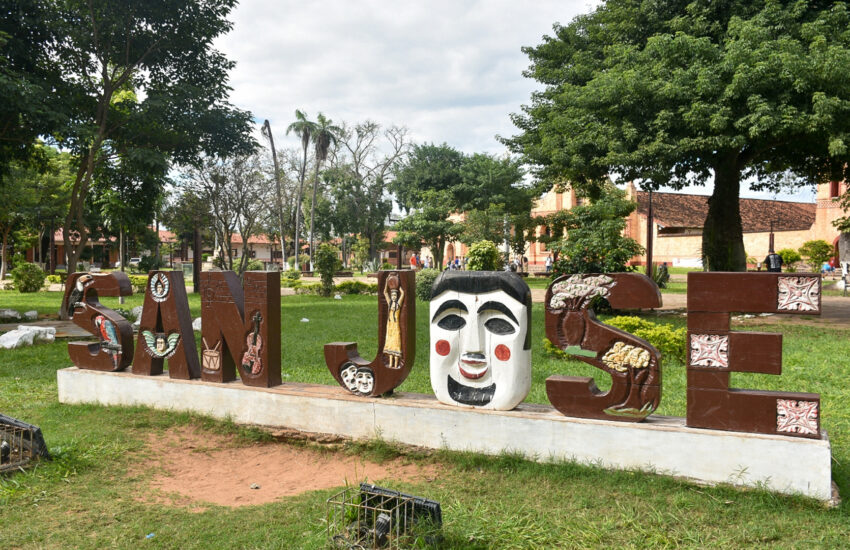 Decorative outdoor sculpture spelling a word with large art-painted letters in a park; each letter features masks and figures, with trees and brick buildings in the background.