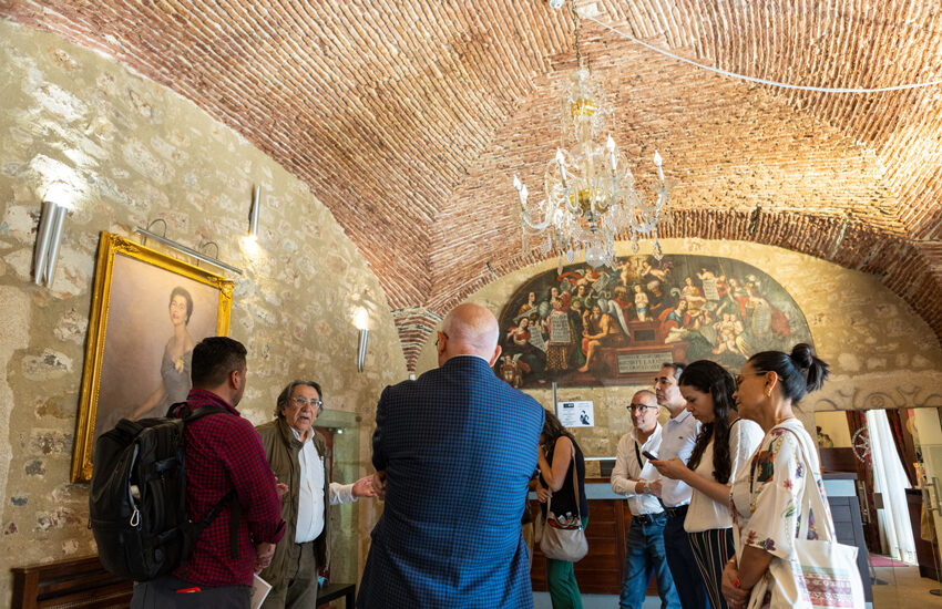 Tour group listening to a guide in a stone-walled room with a mural and chandelier in a historic building.