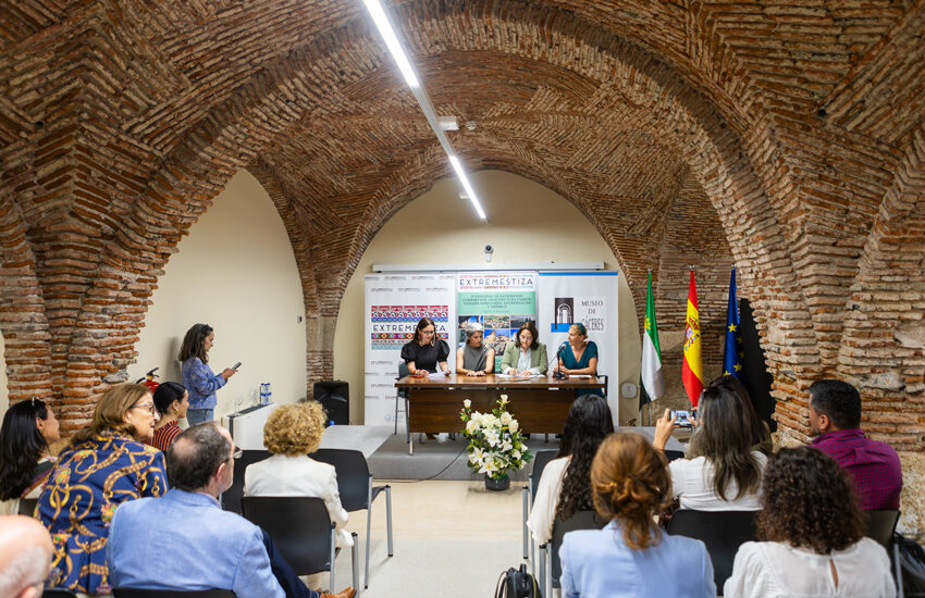 Panel discussion in a brick-arched hall with five speakers at a table, audience listening, flags and banners behind.