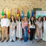 Group of ten people standing in a row for an event, with flags and a stone wall backdrop behind them.