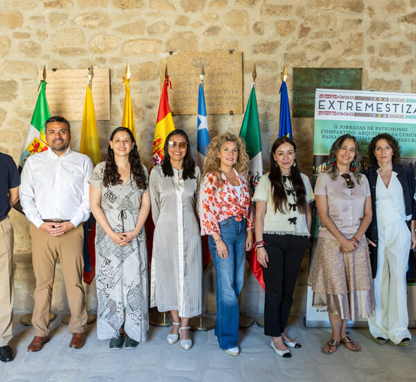 Group of ten people standing in a row for an event, with flags and a stone wall backdrop behind them.