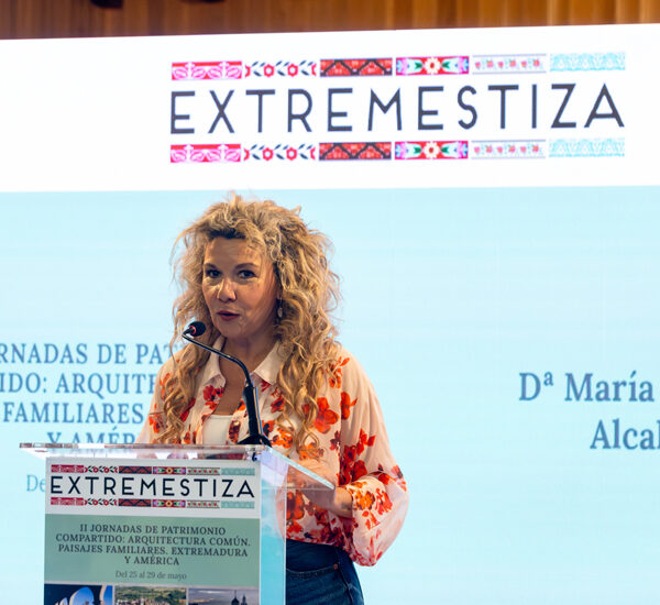 Speaker with curly blonde hair giving a talk at a podium with an EXTREMESTIZA banner and embroidered patterns above.