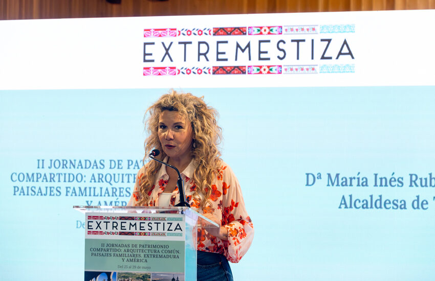 Speaker with curly blonde hair giving a talk at a podium with an EXTREMESTIZA banner and embroidered patterns above.