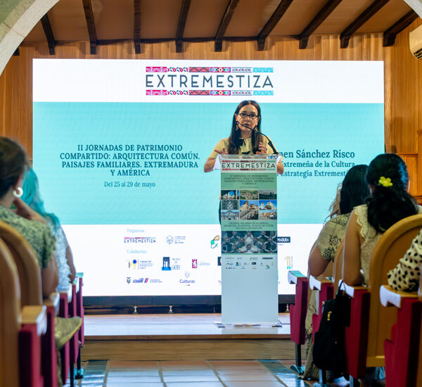 Woman speaks at a conference podium on a stage with a large EXTREMESTIZA banner and blue screen behind her; audience seated in a hall.