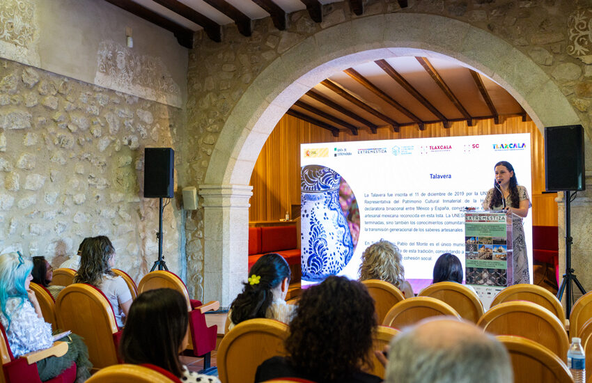 Woman giving a presentation on a stage with a large blue ceramic vase image on the screen behind her, in a stone-walled room with wooden beams and arched doorway.