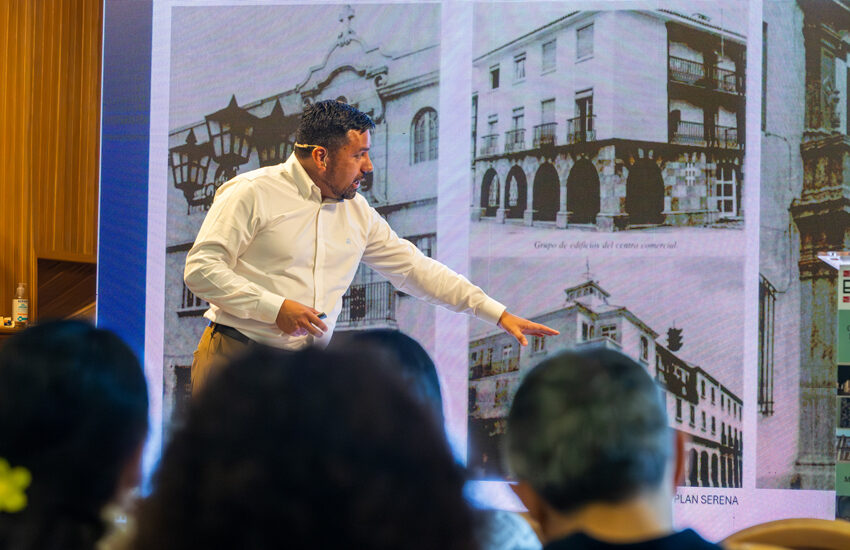 Man in a white shirt giving a presentation on stage, pointing at a large screen displaying black-and-white photos of buildings; audience silhouettes in the foreground.