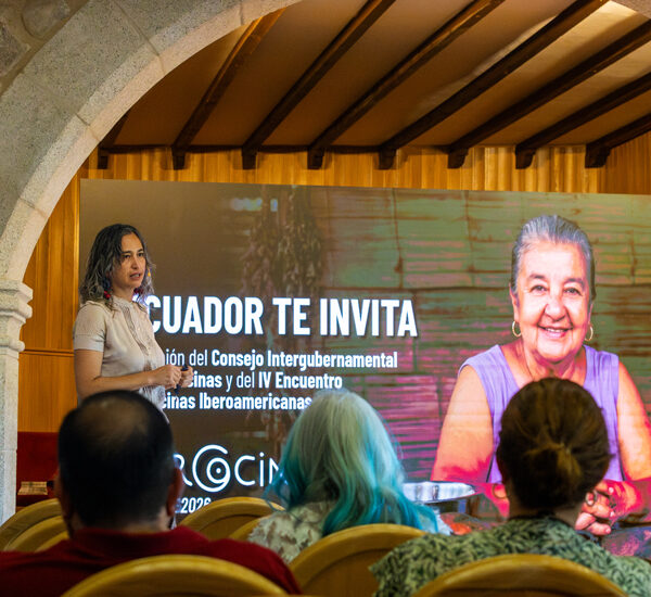 Presenter speaks to an audience in a hall with a large screen that reads 'ECUADOR TE INVITA' and shows a smiling older woman on the right side.
