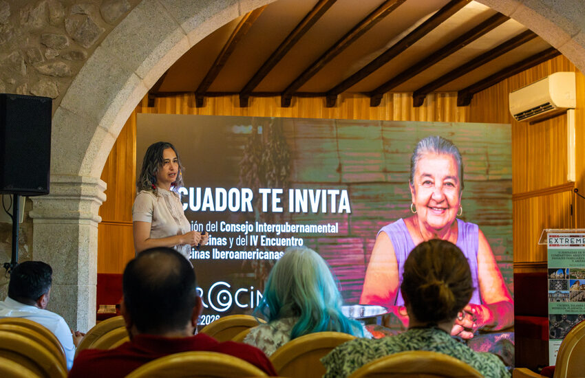 Presenter speaks to an audience in a hall with a large screen that reads 'ECUADOR TE INVITA' and shows a smiling older woman on the right side.