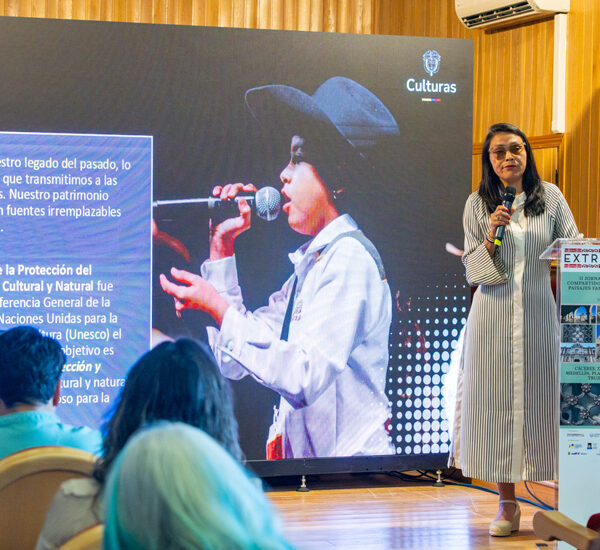 Woman in a striped dress speaks into a microphone on a stage beside a large screen with UNESCO logo and text panel.