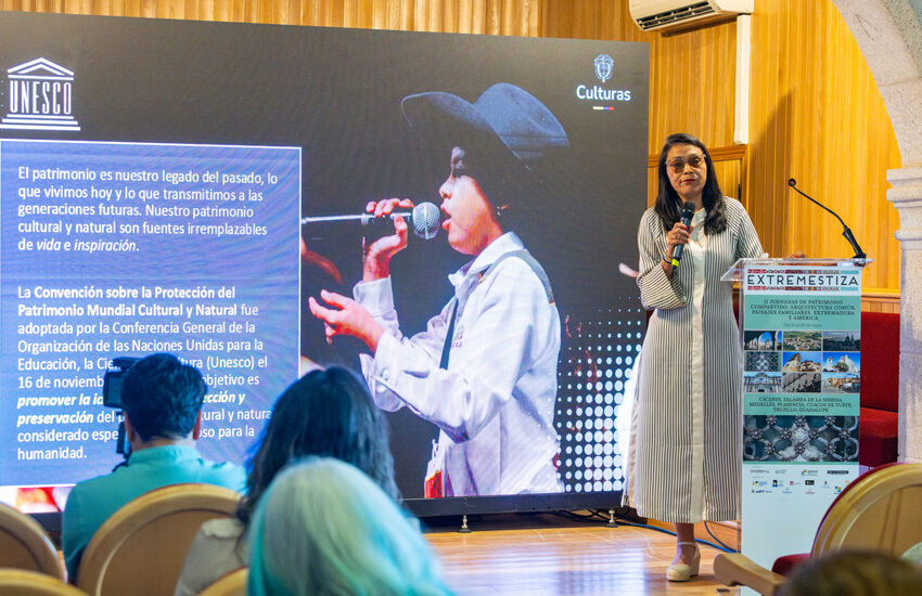 Woman in a striped dress speaks into a microphone on a stage beside a large screen with UNESCO logo and text panel.