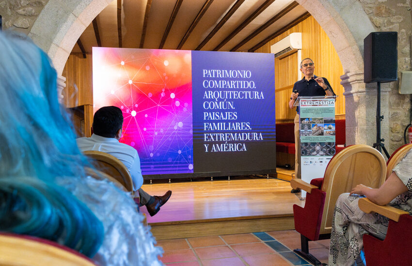 Conference presentation in a stone-walled room; speaker at a podium beside a colorful banner with Spanish text about Extremadura and America fonts visible on stage.