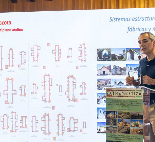 Speaker presenting beside a large screen with architectural diagrams and a poster labeled EXTREMESTIZA at a conference.