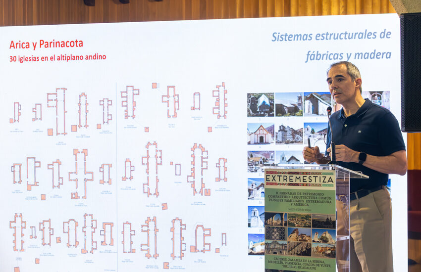 Speaker presenting beside a large screen with architectural diagrams and a poster labeled EXTREMESTIZA at a conference.