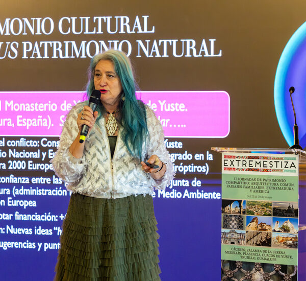Mujer con cabello azul-verdoso habla en micrófono frente a una gran pantalla con el título 'Patrimonio Cultural versus Patrimonio Natural' y un podio a un lado.