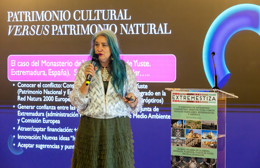 Mujer con cabello azul-verdoso habla en micrófono frente a una gran pantalla con el título 'Patrimonio Cultural versus Patrimonio Natural' y un podio a un lado.