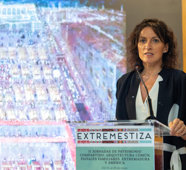 Female speaker at a podium during a conference; the podium banner reads EXTREMESTIZA and mentions II Jornadas de Patrimonio Compartido (architecture, landscapes, Extremadura).
