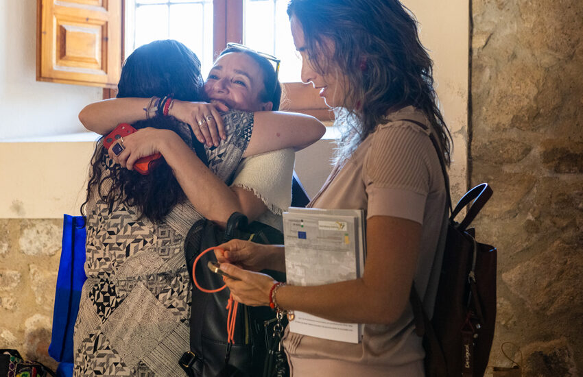 Two women share a warm hug as a third smiling woman holds a clipboard and bag nearby in a sunlit, stone-walled room.