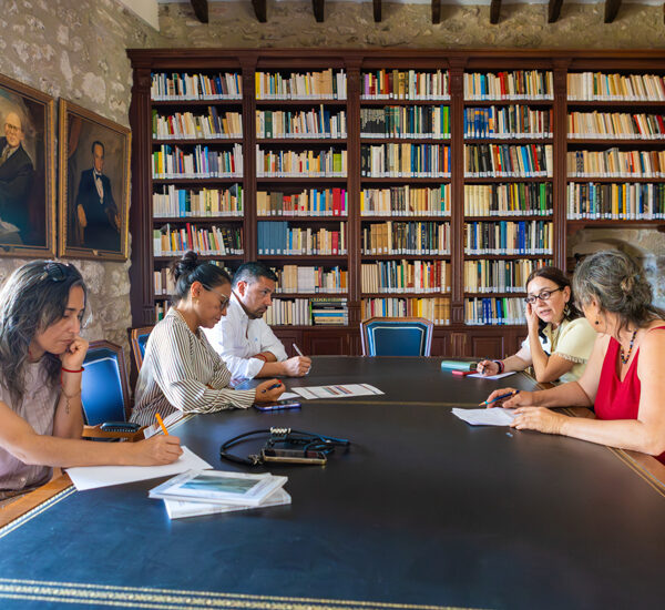 Five people sit around a large oval table in a library, discussing notes with papers and pens on the table.
