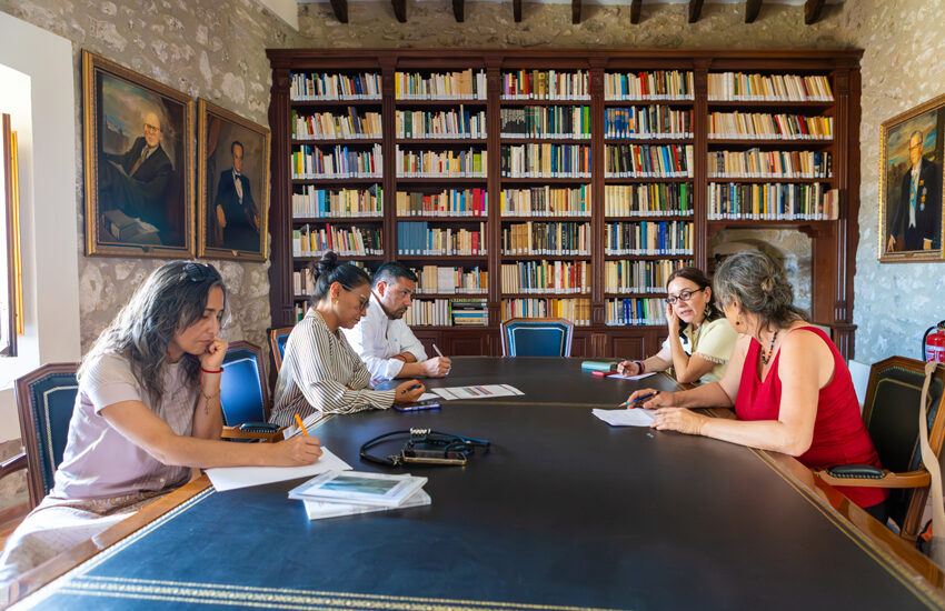 Five people sit around a large oval table in a library, discussing notes with papers and pens on the table.