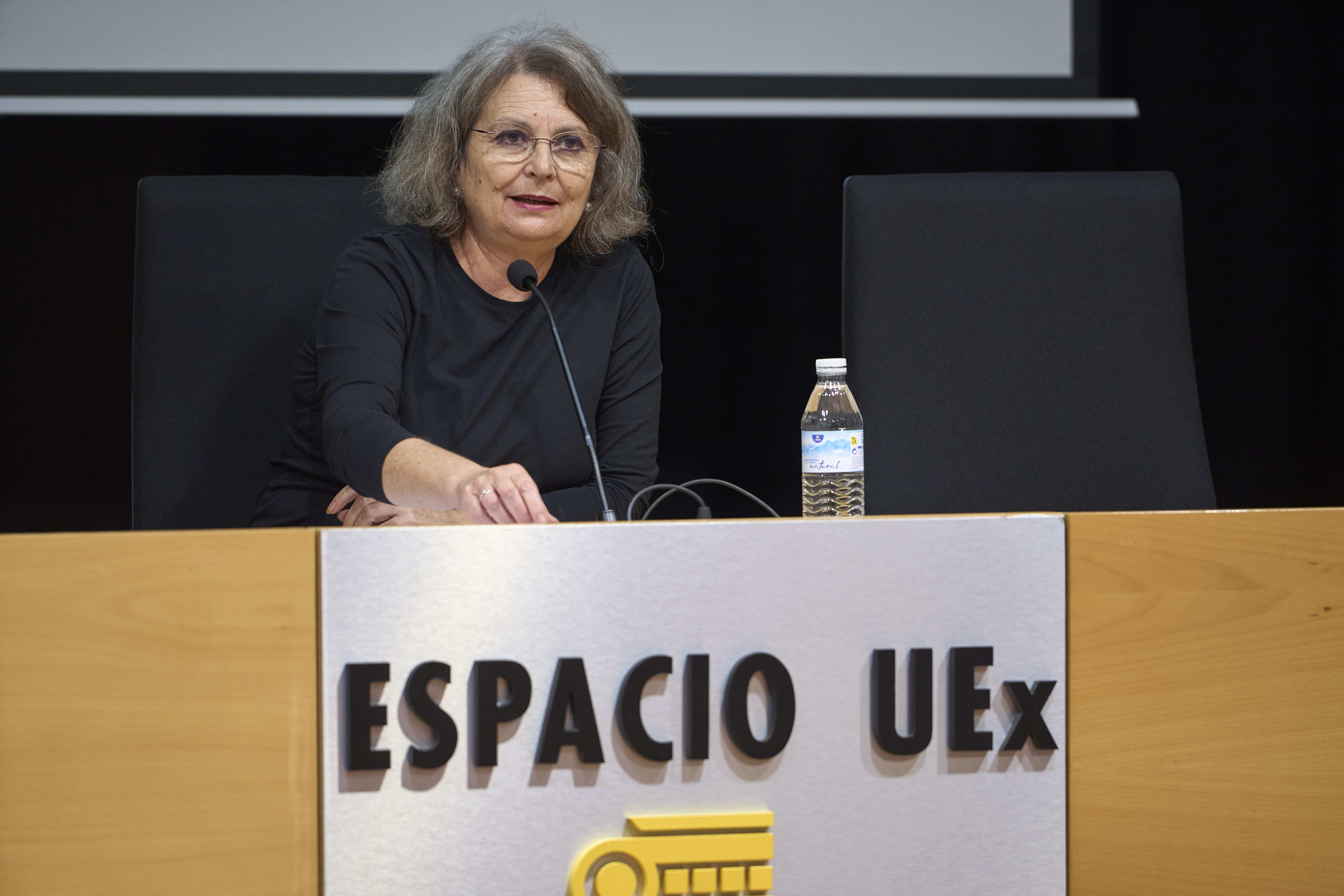 Woman speaking at a conference panel, seated behind a wooden podium with a microphone and water bottle in view.